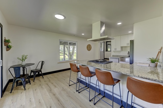 kitchen with a breakfast bar, island range hood, white cabinetry, stainless steel appliances, and light stone countertops