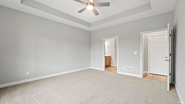 unfurnished bedroom with a tray ceiling, ensuite bathroom, ceiling fan, and light colored carpet