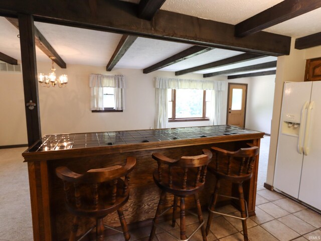 bar featuring white refrigerator with ice dispenser, light tile patterned floors, beam ceiling, an inviting chandelier, and tile countertops