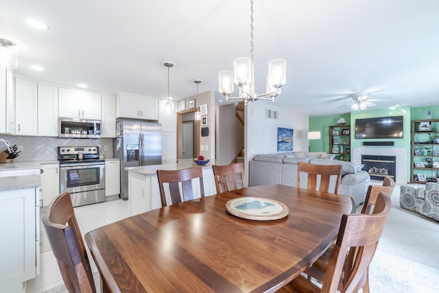 dining area featuring light tile patterned floors, ceiling fan with notable chandelier, a fireplace, visible vents, and stairs