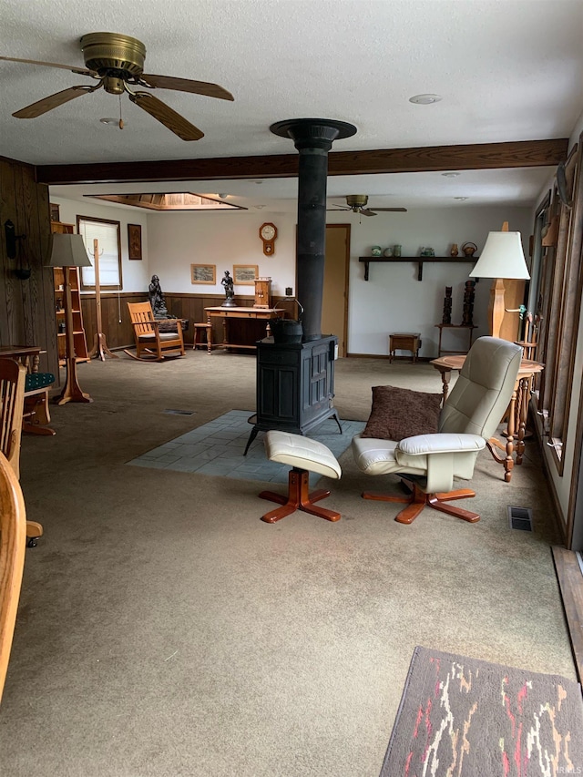 carpeted living room with ceiling fan, a textured ceiling, beam ceiling, and a wood stove