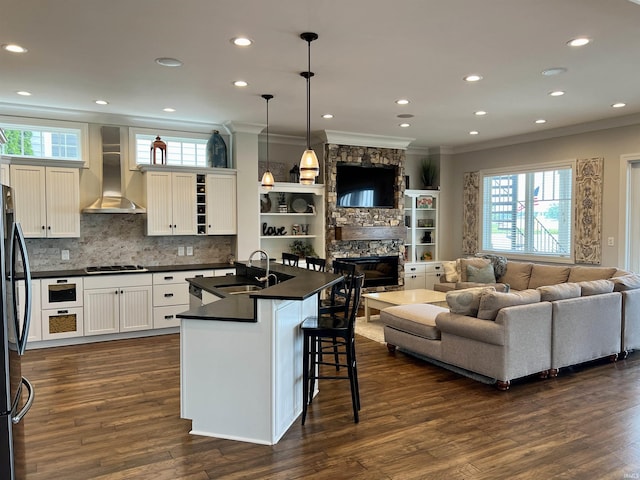 kitchen with wall chimney exhaust hood, sink, a center island with sink, white cabinets, and hanging light fixtures