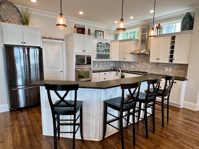 kitchen with wall chimney exhaust hood, hanging light fixtures, an island with sink, and stainless steel appliances