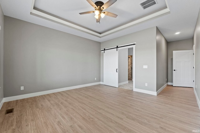 unfurnished bedroom with a tray ceiling, a barn door, ceiling fan, and light wood-type flooring