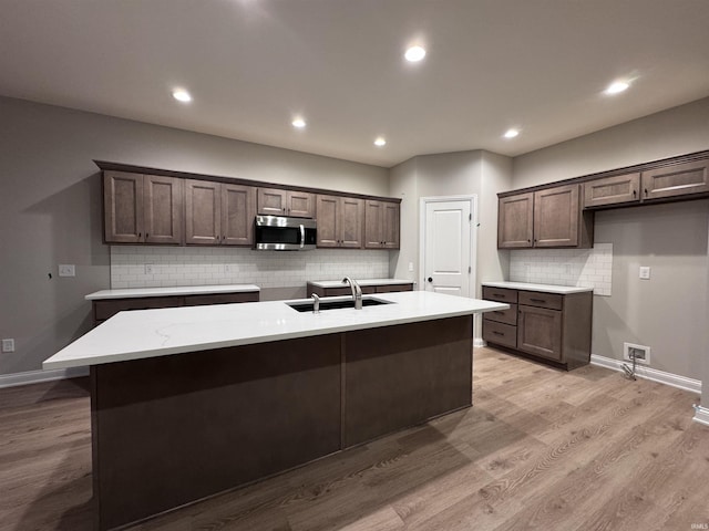 kitchen featuring backsplash, dark brown cabinetry, sink, a center island with sink, and light hardwood / wood-style flooring