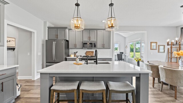 kitchen with gray cabinets, backsplash, an island with sink, stainless steel appliances, and light wood-type flooring