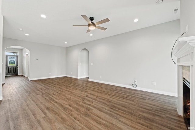 unfurnished living room featuring arched walkways, ceiling fan, wood finished floors, a lit fireplace, and baseboards