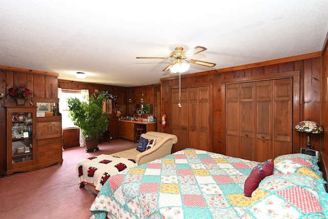 carpeted bedroom featuring wood walls, two closets, ceiling fan, and a textured ceiling