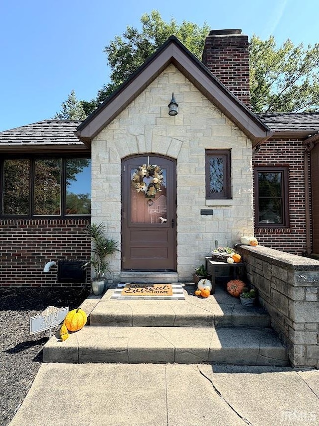 doorway to property featuring brick siding, stone siding, a chimney, and roof with shingles