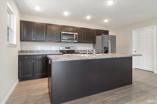 kitchen with appliances with stainless steel finishes, a kitchen island with sink, light countertops, light wood-type flooring, and a sink