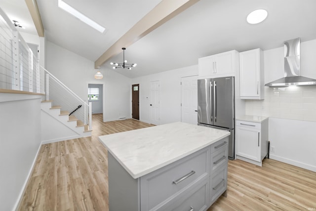 kitchen featuring wall chimney range hood, light wood-type flooring, backsplash, stainless steel refrigerator, and lofted ceiling with beams