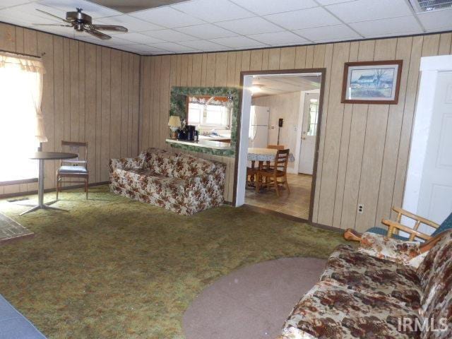 carpeted living room with ceiling fan, wooden walls, and a drop ceiling