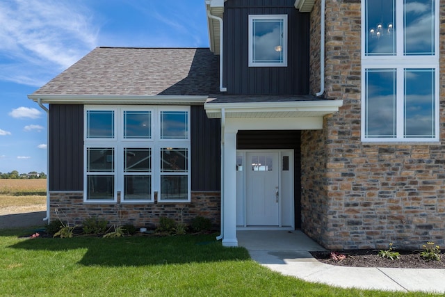 view of exterior entry with board and batten siding, stone siding, a yard, and a shingled roof