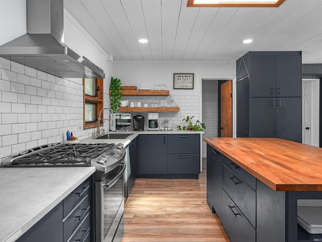 kitchen with stainless steel gas range, wooden counters, open shelves, a sink, and wall chimney exhaust hood