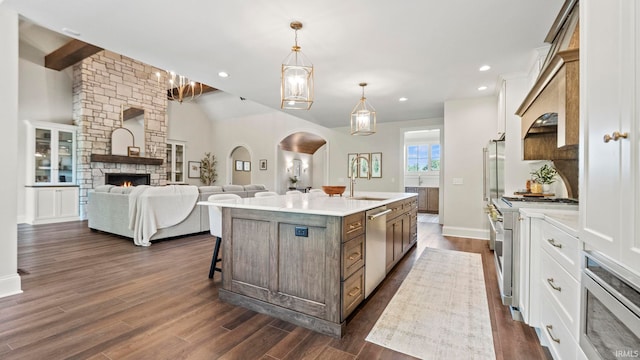 kitchen with stainless steel appliances, decorative light fixtures, an island with sink, dark wood-type flooring, and a stone fireplace