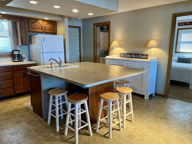 kitchen featuring stone tile flooring, freestanding refrigerator, a kitchen island with sink, a sink, and a kitchen breakfast bar