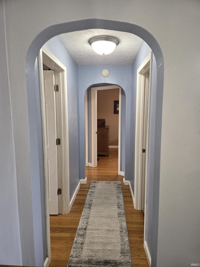 hall with a textured ceiling, baseboards, arched walkways, and dark wood-style flooring