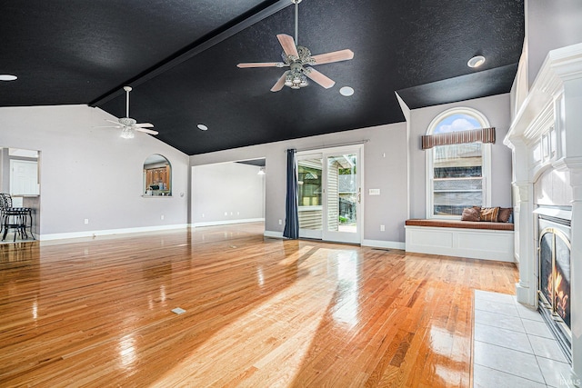 unfurnished living room featuring ceiling fan, beam ceiling, light hardwood / wood-style flooring, high vaulted ceiling, and a tile fireplace