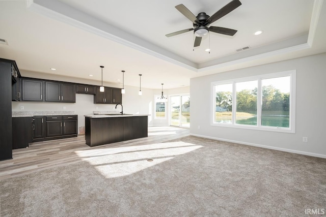 kitchen with pendant lighting, light wood-type flooring, a tray ceiling, ceiling fan, and a kitchen island with sink