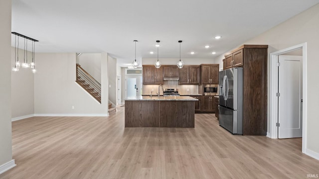 kitchen with light wood-type flooring, an island with sink, appliances with stainless steel finishes, decorative light fixtures, and light stone countertops