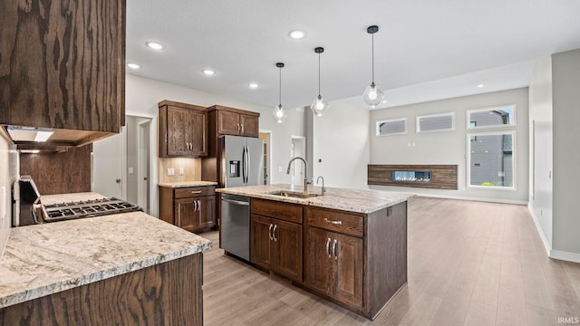 kitchen with light stone counters, a kitchen island with sink, sink, hanging light fixtures, and appliances with stainless steel finishes