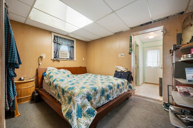 carpeted bedroom with a drop ceiling, cooling unit, wooden walls, and multiple windows