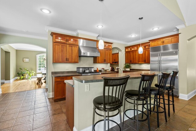 kitchen featuring pendant lighting, an island with sink, wall chimney range hood, appliances with stainless steel finishes, and a breakfast bar area