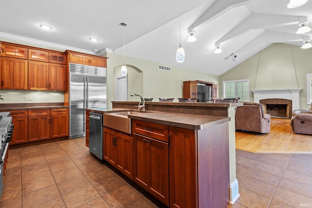 kitchen featuring a tile fireplace, vaulted ceiling, hanging light fixtures, a center island with sink, and appliances with stainless steel finishes