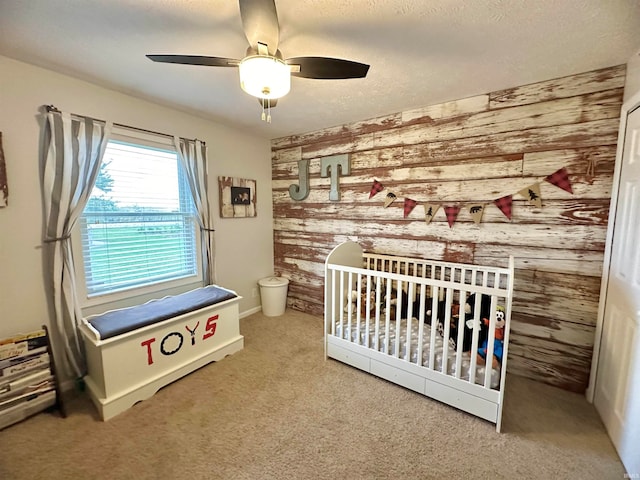 carpeted bedroom featuring ceiling fan, a textured ceiling, wooden walls, and a crib