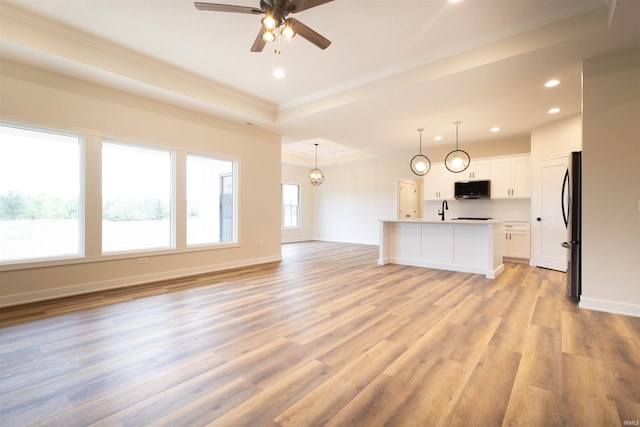 unfurnished living room with light wood-type flooring, a tray ceiling, sink, ornamental molding, and ceiling fan