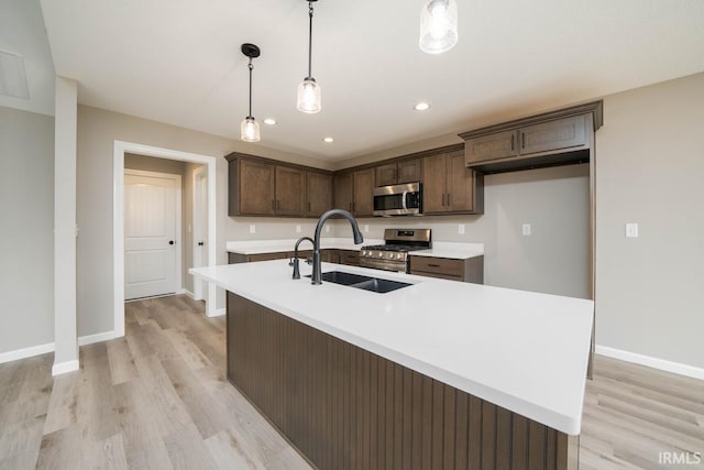 kitchen with sink, pendant lighting, a kitchen island with sink, appliances with stainless steel finishes, and light wood-type flooring