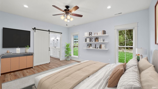 bedroom with a barn door, connected bathroom, light hardwood / wood-style floors, and ceiling fan