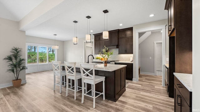 kitchen featuring pendant lighting, dark brown cabinets, light hardwood / wood-style floors, backsplash, and a center island with sink