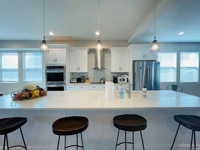 kitchen featuring white cabinets, appliances with stainless steel finishes, hanging light fixtures, and wall chimney range hood