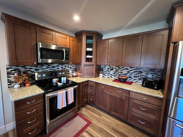 kitchen with dark brown cabinets, light hardwood / wood-style floors, decorative backsplash, appliances with stainless steel finishes, and light stone countertops