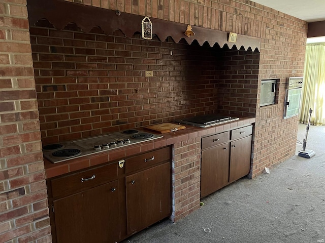 kitchen with stainless steel gas stovetop, brick wall, light colored carpet, and dark brown cabinetry