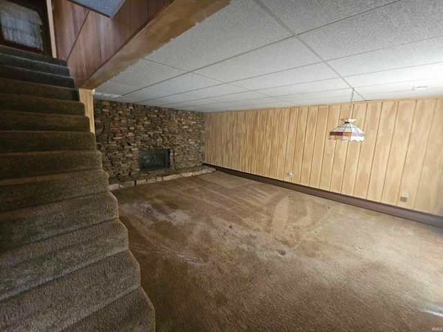 basement with a stone fireplace, a drop ceiling, carpet floors, and wood walls