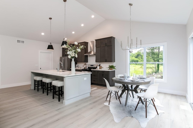 kitchen featuring light wood-type flooring, an island with sink, stainless steel appliances, wall chimney exhaust hood, and pendant lighting