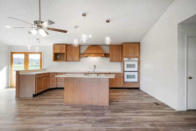 kitchen with an island with sink, custom range hood, white double oven, and dark hardwood / wood-style flooring