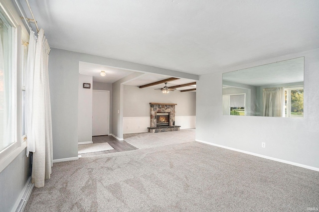 unfurnished living room with beamed ceiling, a stone fireplace, light colored carpet, and ceiling fan