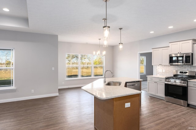 kitchen featuring an island with sink, hanging light fixtures, sink, appliances with stainless steel finishes, and light stone counters