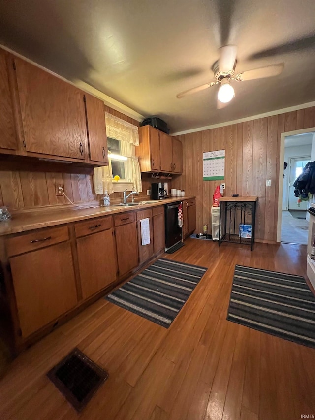 kitchen featuring sink, dishwasher, light hardwood / wood-style floors, ceiling fan, and wooden walls