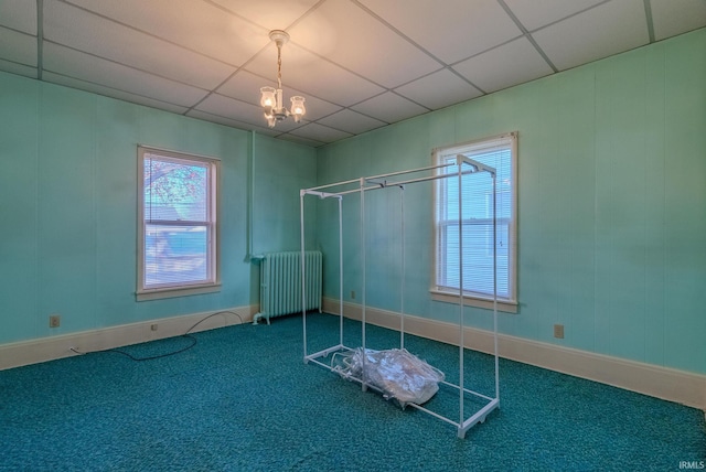 carpeted empty room featuring a drop ceiling, radiator heating unit, and a notable chandelier