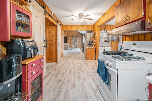 kitchen with wood walls, white gas range, light hardwood / wood-style flooring, a barn door, and ceiling fan