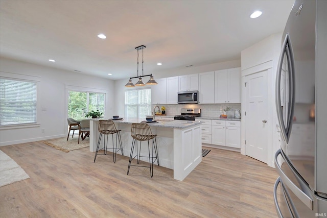 kitchen with an island with sink, stainless steel appliances, light stone countertops, decorative light fixtures, and white cabinetry