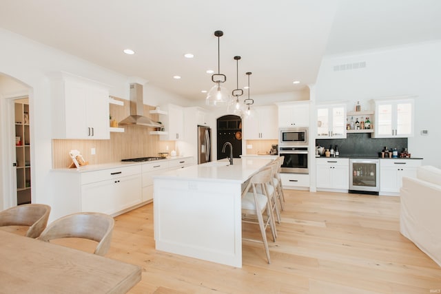 kitchen featuring wall chimney range hood, appliances with stainless steel finishes, hanging light fixtures, wine cooler, and a kitchen island with sink