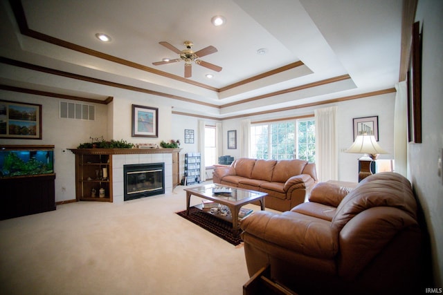 carpeted living room featuring a tiled fireplace, a raised ceiling, crown molding, and ceiling fan