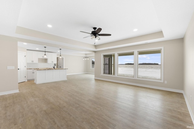 unfurnished living room featuring ceiling fan with notable chandelier, a tray ceiling, light hardwood / wood-style flooring, and sink