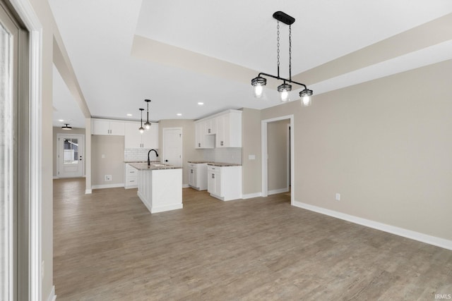 kitchen with decorative backsplash, light wood-type flooring, an island with sink, decorative light fixtures, and white cabinetry