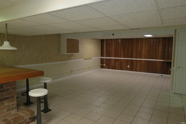 basement with wood walls, light tile patterned floors, electric panel, and a drop ceiling